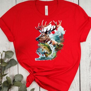 May include: A red t-shirt with a detailed graphic of a deer with large antlers, a fish, and a mountain scene. The design incorporates shades of brown, green, and blue. The shirt is made of a soft material.