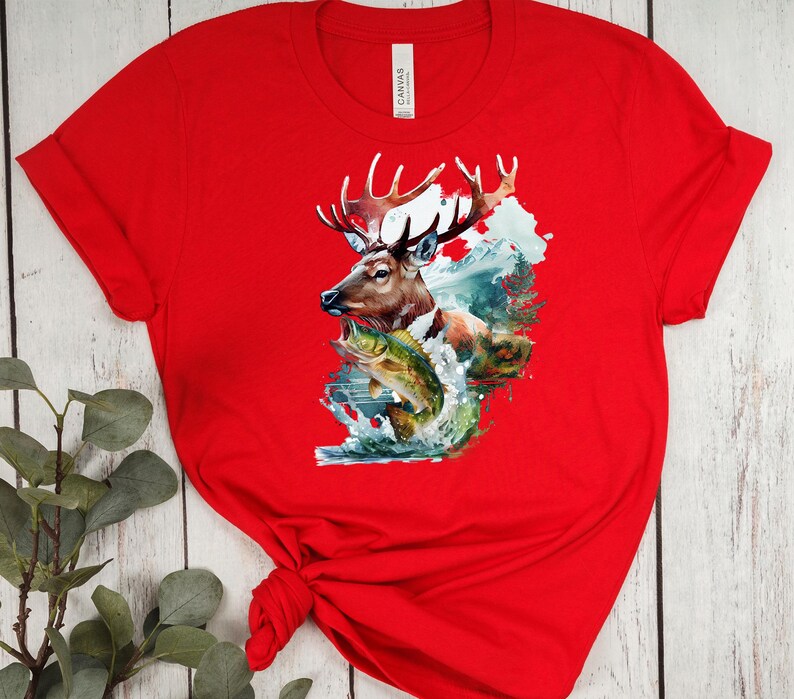 May include: A red t-shirt with a detailed graphic of a deer with large antlers, a fish, and a mountain scene. The design incorporates shades of brown, green, and blue. The shirt is made of a soft material.