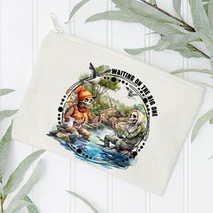 K&ouml;nnte beinhalten: Eine wei&szlig;e Canvas-Kosmetiktasche mit einer Schwarzwei&szlig;illustration von zwei Skeletten beim Angeln. Der Text "Waiting on the Big One" ist auf der Tasche aufgedruckt.