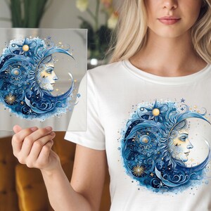 May include: A white t-shirt with a blue and gold crescent moon design. The moon is adorned with intricate floral details and stars. The design features a stylized face with a golden halo.