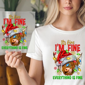 May include: A clear square with a graphic of a Highland cow wearing a Santa hat and Christmas lights. Text reads "It's fine, I'm fine, Everything is fine" in red and green. The cow has brown fur and is sticking out its tongue.