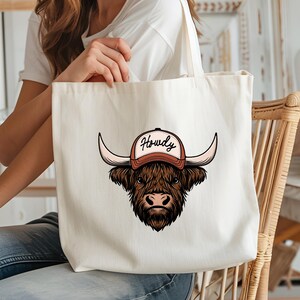 May include: A cream-colored tote bag featuring a brown Highland cow wearing a baseball cap with the word "Howdy" in a script font. The cow has large horns and a shaggy coat. The bag has long straps.