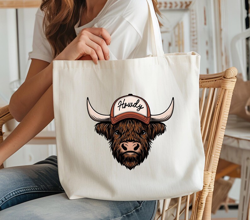 May include: A cream-colored tote bag featuring a brown Highland cow wearing a baseball cap with the word "Howdy" in a script font. The cow has large horns and a shaggy coat. The bag has long straps.