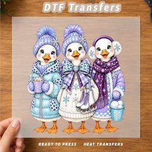 May include: A DTF transfer sheet featuring three cartoon geese dressed for winter. The left goose wears a blue puffer coat and hat, holding a cocoa mug. The center goose wears a white sweater and purple bow, holding ice skates. The right goose wears a purple scarf and earmuffs, holding a bucket of snowballs.