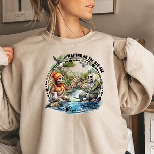 K&ouml;nnte beinhalten: Ein beiger Pullover mit einem grafischen Aufdruck von zwei Skeletten beim Angeln. Der Text auf dem Aufdruck lautet "Waiting on the big one".