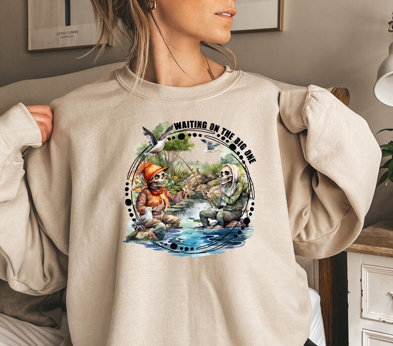 K&ouml;nnte beinhalten: Ein beiger Pullover mit einem grafischen Aufdruck von zwei Skeletten beim Angeln. Der Text auf dem Aufdruck lautet "Waiting on the big one".