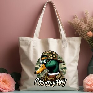May include: A white canvas tote bag featuring a cartoon duck wearing a camouflage hat and sunglasses. The duck is sporting a gold chain and the text "Country Boy" is printed beneath it.