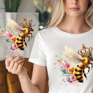 May include: A watercolor illustration of a bee wearing a golden crown, with yellow and black stripes, and delicate wings. The bee is surrounded by colorful flowers in shades of pink, purple, and yellow. The design is on a clear square and a white t-shirt.