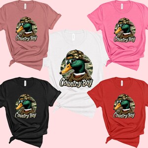 May include: Five t-shirts in different colours featuring a cartoon duck wearing a backwards baseball cap and sunglasses. The duck is wearing a gold chain and the text "Country Boy" is printed below the duck. The t-shirts are available in pink, red, black, white, and brown.