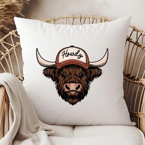 May include: White throw pillow featuring a cartoon illustration of a Highland cow wearing a brown and white baseball cap. The cap has the word "Howdy" in black script. The cow has long brown fur and large horns.