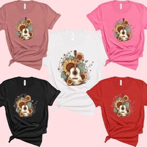 May include: Five different color t-shirts featuring a graphic of an acoustic guitar with sunflowers. The guitar is brown with a white pickguard and the sunflowers are yellow with green leaves.