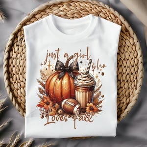 May include: White t-shirt with a graphic design of a pumpkin, a cup of coffee, and a football. The text "just a girl who loves fall" is printed on the shirt.