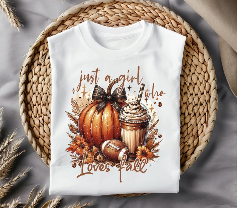 May include: White t-shirt with a graphic design of a pumpkin, a cup of coffee, and a football. The text "just a girl who loves fall" is printed on the shirt.