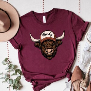 May include: Burgundy t-shirt featuring a detailed illustration of a Highland cow wearing a baseball cap with the word "Howdy" on it. The cow has long horns and a shaggy brown coat. The shirt is a solid color.