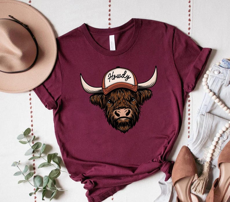 May include: Burgundy t-shirt featuring a detailed illustration of a Highland cow wearing a baseball cap with the word "Howdy" on it. The cow has long horns and a shaggy brown coat. The shirt is a solid color.