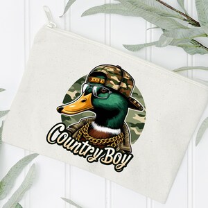 May include: A white canvas makeup bag featuring a cartoon illustration of a duck wearing a camouflage hat and sunglasses. The duck has a gold chain around its neck and the text "Country Boy" is printed below the duck.