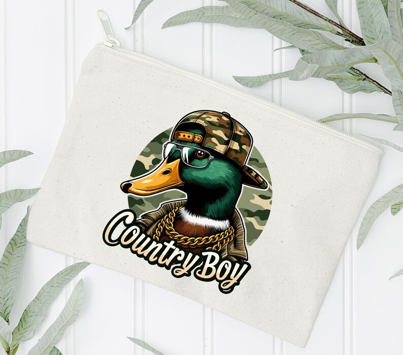 May include: A white canvas makeup bag featuring a cartoon illustration of a duck wearing a camouflage hat and sunglasses. The duck has a gold chain around its neck and the text "Country Boy" is printed below the duck.