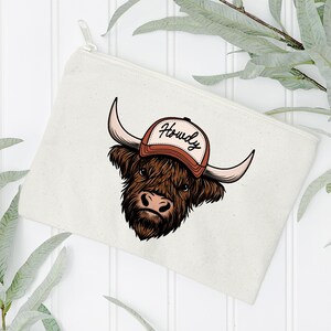 May include: A cream-colored zippered pouch featuring a cartoon illustration of a Highland cow wearing a brown and white baseball cap with the word "Howdy". The cow has brown fur, large horns, and a pink nose.