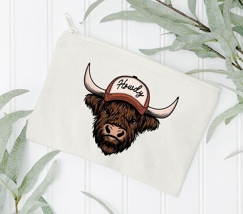 May include: A cream-colored zippered pouch featuring a cartoon illustration of a Highland cow wearing a brown and white baseball cap with the word "Howdy". The cow has brown fur, large horns, and a pink nose.