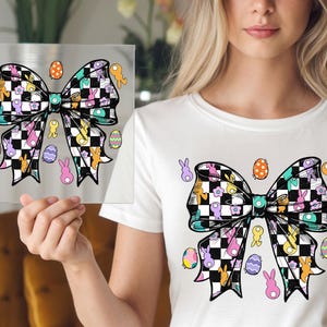 May include: A white t-shirt with a black and white checkered bow with pastel Easter eggs and bunnies. The bow is centered on the shirt.