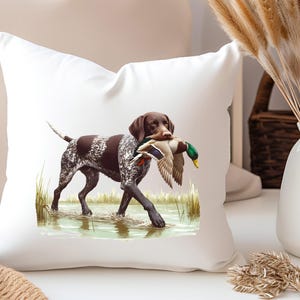 Puede incluir: Una almohada blanca con un perro Braco Alem&aacute;n de Pelo Corto marr&oacute;n y blanco que lleva un pato silvestre verde y marr&oacute;n en la boca. El perro est&aacute; de pie en una zona herbosa con agua.