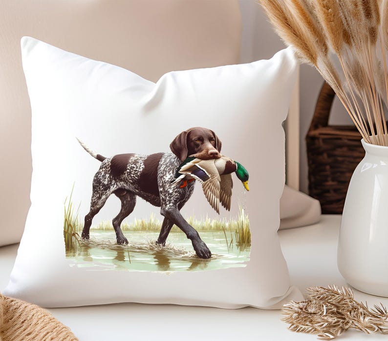 Puede incluir: Una almohada blanca con un perro Braco Alem&aacute;n de Pelo Corto marr&oacute;n y blanco que lleva un pato silvestre verde y marr&oacute;n en la boca. El perro est&aacute; de pie en una zona herbosa con agua.