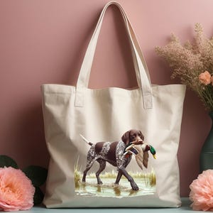 Puede incluir: Una bolsa de lona blanca con un perro Braco Alem&aacute;n de Pelo Corto marr&oacute;n y blanco que lleva un pato silvestre en la boca. El perro est&aacute; de pie en una zona herbosa con agua.