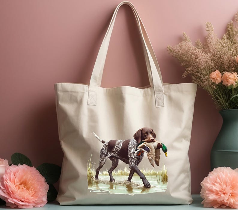 Puede incluir: Una bolsa de lona blanca con un perro Braco Alem&aacute;n de Pelo Corto marr&oacute;n y blanco que lleva un pato silvestre en la boca. El perro est&aacute; de pie en una zona herbosa con agua.