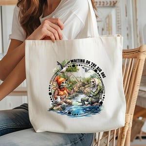 K&ouml;nnte beinhalten: Eine wei&szlig;e Canvas-Tragetasche mit einer Schwarzwei&szlig;-Illustration von zwei Skeletten beim Angeln. Der Text "Waiting on the big one" ist in Schwarz auf die Tasche gedruckt.