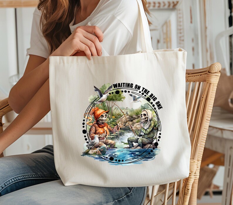 K&ouml;nnte beinhalten: Eine wei&szlig;e Canvas-Tragetasche mit einer Schwarzwei&szlig;-Illustration von zwei Skeletten beim Angeln. Der Text "Waiting on the big one" ist in Schwarz auf die Tasche gedruckt.