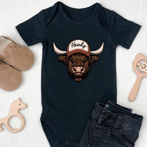 May include: Black baby bodysuit with a graphic of a Highland cow wearing a cap that says "Howdy". The image includes a pair of brown baby shoes, a wooden horse-shaped teether, a wooden rattle, and a pair of dark denim jeans.