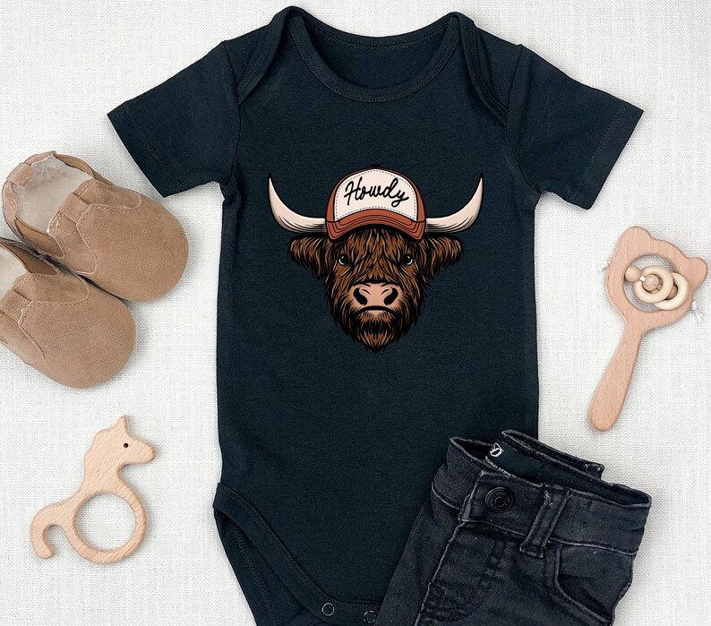 May include: Black baby bodysuit with a graphic of a Highland cow wearing a cap that says "Howdy". The image includes a pair of brown baby shoes, a wooden horse-shaped teether, a wooden rattle, and a pair of dark denim jeans.