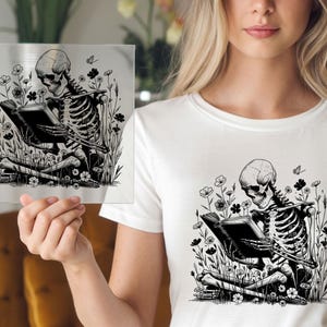 May include: Black and white illustration of a skeleton reading a book while sitting in a field of flowers. The artwork is on a clear square, held in front of a white t-shirt with the same design. The theme is gothic and whimsical.