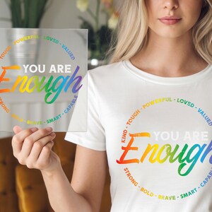 May include: A clear square glass with the words "YOU ARE Enough" in rainbow colors. The words "Kind, Tough, Powerful, Loved, Valued, Capable, Smart, Brave, Bold, Strong" are in a circle around the main text. A white t-shirt with the same design is also visible.