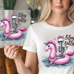 May include: A white t-shirt with a pink and black skeleton riding a pink unicorn float with the text "Stay Salty".