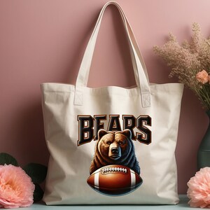 May include: A white canvas tote bag with brown straps. The bag has a brown bear holding a football with the word "BEARS" in brown letters above the bear.