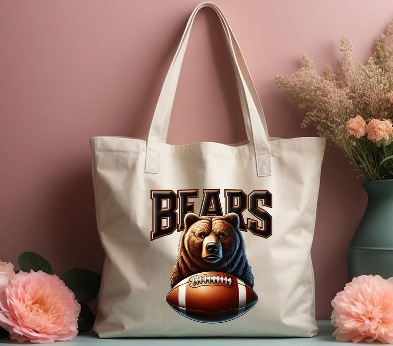 May include: A white canvas tote bag with brown straps. The bag has a brown bear holding a football with the word "BEARS" in brown letters above the bear.