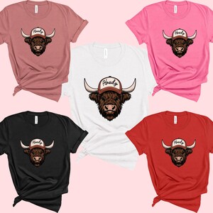 May include: A collection of t-shirts in various colors, including pink, red, black, white, and mauve. Each shirt features a graphic of a Highland cow wearing a baseball cap with the word "Howdy" on it.