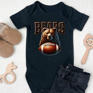 May include: A black baby onesie with a brown bear wearing a football helmet and holding a football. The onesie has the word "BEARS" printed on it in orange letters.