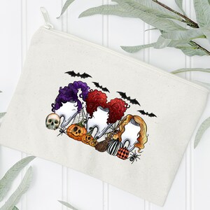 May include: A white canvas zippered pouch with a Halloween-themed design featuring three witches with their hair in a bun, wearing a tooth as a hat. The witches are surrounded by bats, pumpkins, and a skull.