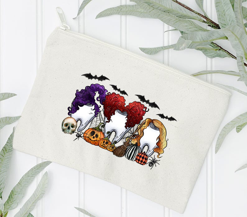 May include: A white canvas zippered pouch with a Halloween-themed design featuring three witches with their hair in a bun, wearing a tooth as a hat. The witches are surrounded by bats, pumpkins, and a skull.