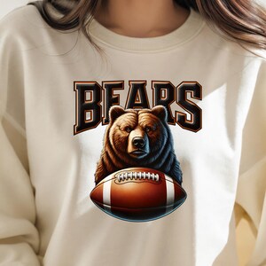 May include: A cream-colored sweatshirt with a brown bear holding a football. The text "BEARS" is printed in black above the bear.