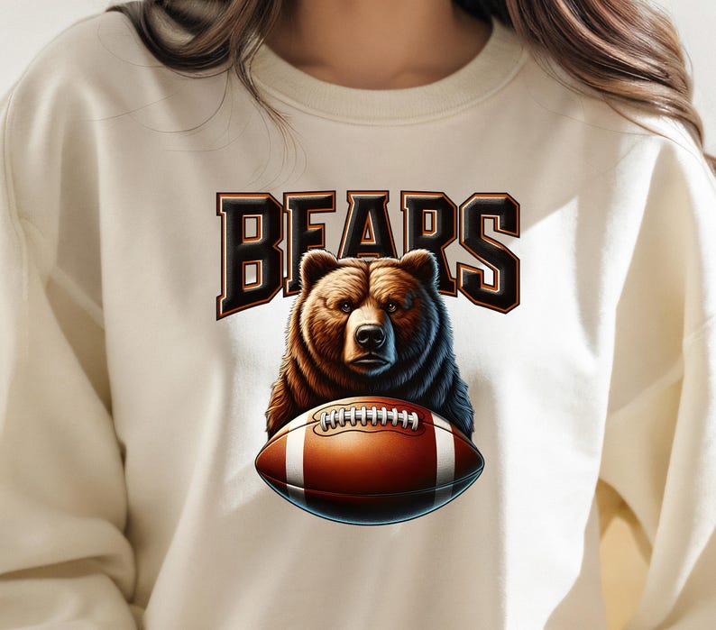 May include: A cream-colored sweatshirt with a brown bear holding a football. The text "BEARS" is printed in black above the bear.