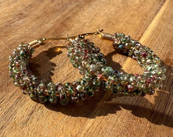 May include: A pair of hoop earrings with a gold-coloured metal base. The hoops are decorated with small, colourful beads in shades of green, red, and clear, creating a textured look. The earrings are displayed on a wooden surface.