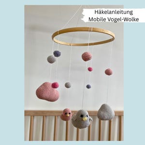 Può includere: Mobile per neonati fatto a mano con anello in legno ed elementi all'uncinetto sospesi. Include uccelli, nuvole e sfere rosa, grigie e viola. Il testo "Häkelanleitung Mobile Vogel-Wolke" è visibile.