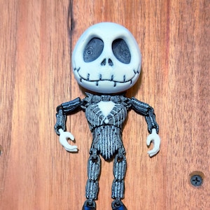 May include: A black and white articulated toy figure of Jack Skellington from the Nightmare Before Christmas movie. The figure has a black and white striped suit and a black bow tie. The figure is posed with its arms outstretched and its legs slightly apart.