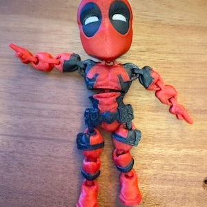 May include: A red and black articulated action figure of Deadpool. The figure is posed with one arm outstretched and the other bent at the elbow. The figure has a large head with a red mask and black eyes.