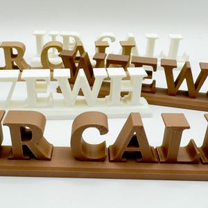 May include: Brown and white 3D-printed letter signs spelling out "UR CALL" and "WE WH" on a white surface. The letters are in a bold, sans-serif font. The signs are decorative and suitable for home or office decor.