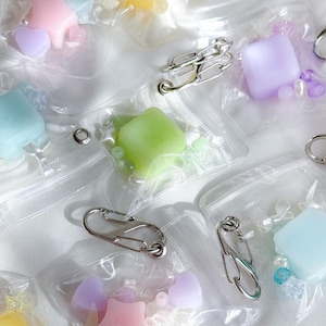 May include: A collection of small, clear plastic bags, each containing colorful, pastel-colored shapes like stars, hearts, and squares, along with small beads. The bags are secured with silver-colored safety pins and carabiner clips.