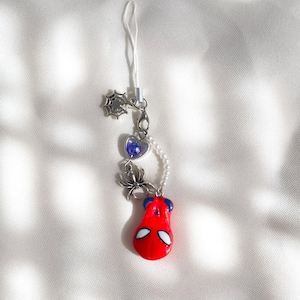 Puede incluir: Un colgante para teléfono con una máscara de superhéroe roja y azul, un dije de araña plateado, un dije de telaraña plateado, un pequeño dije de corazón morado y una hilera de perlas blancas falsas unidas a un cordón blanco para colgar.
